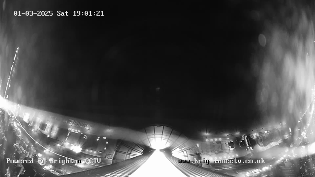 A black and white image from a webcam showing a view of city streets at night. The scene appears dark with some illuminated areas, likely from streetlights and vehicle headlights. In the center, there is a bright light source, possibly from an overhead structure. The date and time are displayed at the top, indicating it is March 1, 2025, at 19:01:21. The bottom left corner includes the text "Powered By Brighton CCTV" and the website URL appears in the bottom right corner.