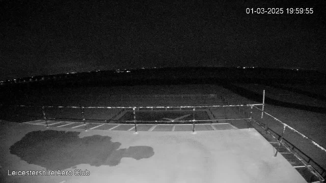 A dark image captured from a webcam overlooking an open area at night. The foreground features a flat surface with some blurred shapes, possibly shadows. In the background, there are distant lights visible, indicating the presence of a town or city beyond the open space. A railing is present along the edge, and the time stamp in the upper right corner shows "01-03-2025 19:59:55." The overall scene has a high contrast between the dark sky and the faint lights.