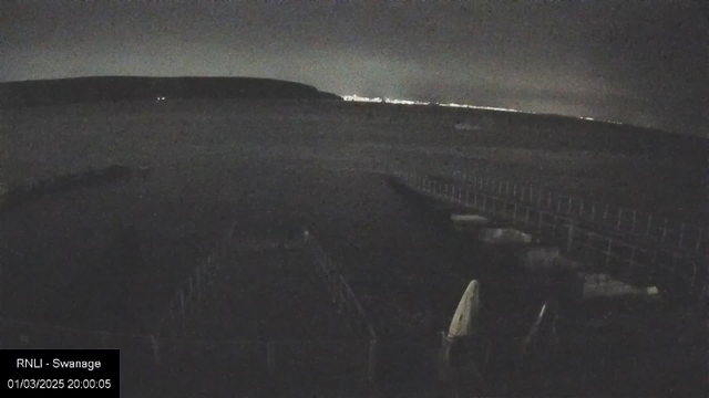 A dark scene showing a calm sea under a night sky. In the foreground, there are outlines of boats and a pier with wooden planks. The horizon features faint lights from a distant shoreline. The atmosphere is serene but dimly lit, suggesting evening or nighttime.