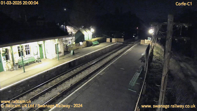 A quiet train station at night, featuring a stone building with green trim and several green benches along the platform. The area is dimly lit with a street lamp and a small wooden fence. Two sets of railway tracks run through the station, and a sign on the platform reads "Way Out". In the background, there are trees and shadows, enhancing the nighttime atmosphere. The scene conveys a sense of calmness and solitude.