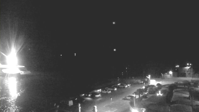 A dark scene captured by a webcam, showing a partially illuminated harbor area at night. The left side features a bright light source, possibly from a boat or a docked vessel, reflecting on the water. Several dim lights are visible along the shore, highlighting a few parked vehicles. The overall atmosphere is subdued with a few distant point lights visible in the dark sky.