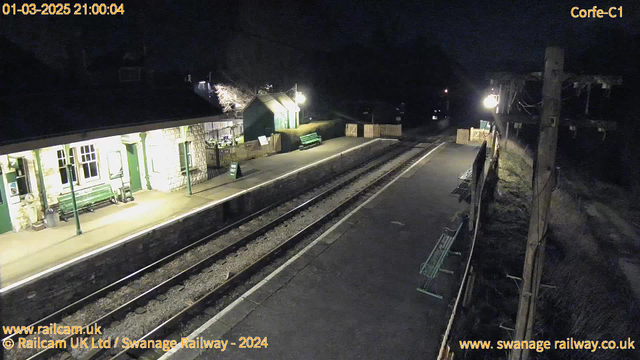 A dark scene of a railway station at night. The platform is mostly empty, featuring several green benches along the edge. To the left, there is a station building with large windows and a sloped roof, illuminated by a soft light. A sign reading "Way Out" is propped against the wall near the entrance. On the right, there is a wooden fence enclosing part of the area, and another green bench is visible. The railway tracks run parallel to the platform, with gravel in between. A faint light from a streetlamp casts a glow on the scene, enhancing the visibility of the surroundings in the dim lighting.