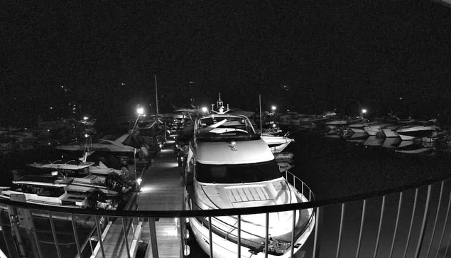 A nighttime image of a marina with numerous boats docked in the water. In the foreground, a large white boat is prominently visible, with its bow facing towards the camera and some details on its deck illuminated. The dock is lined with other boats, partially shadowed, and there are faint lights illuminating the scene, creating a contrast against the dark water and surrounding area. The overall ambiance is calm and quiet, typical of a nighttime setting at a marina.