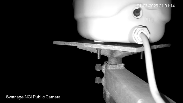 A close-up view of a white container resting on a metal platform. The platform is attached to a metallic pole, with visible screws and a power cable connected to the container. The background is dark, indicating low light conditions. A timestamp in the corner indicates the date and time of the image.