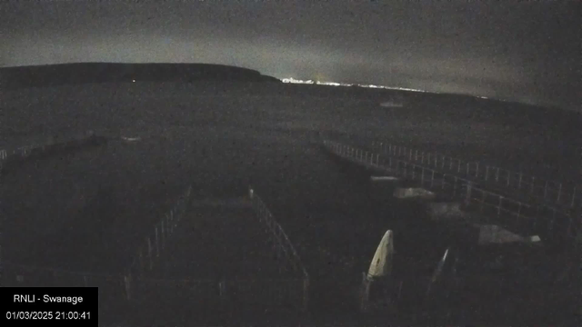 A nighttime scene by the coastline shows dark water with faint light reflections. In the foreground, wooden structures and a few boat covers are visible. A distant hill is silhouetted against the sky, while lights from a faraway location create a subtle glow near the horizon. The overall ambiance is calm and tranquil, but the image is quite dark.