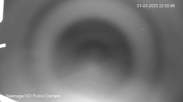 A blurred black and white image of a circular area with a faint gradient, appearing as if looking into a tunnel or camera lens. There are vague outlines and minimal contrast, creating an abstract visual effect. A timestamp is visible in the upper right corner, indicating the date and time. The bottom left section includes text identifying the camera as a public webcam.