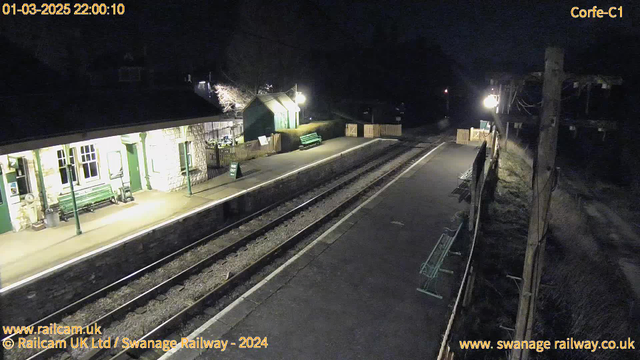 A nighttime image of a railway station with empty platforms. There is a stone building with green accents and large windows on the left, which appears well lit. A green bench is visible in front of the building, and a sign that says "MAN OUT" can be seen nearby. The platform has two sets of railway tracks running parallel, with a wooden fence in the background. On the right side, there’s a lamp post casting light, and another green bench is placed on the platform. The scene is quiet and deserted, emphasizing the darkness surrounding the station.