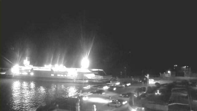 A brightly lit ferry docked at a harbor at night, with reflections shimmering on the water. Surrounding the ferry are multiple parked cars, and streetlights illuminating the area. There are structures and trees in the foreground, while the background is mostly dark, indicating it is nighttime.