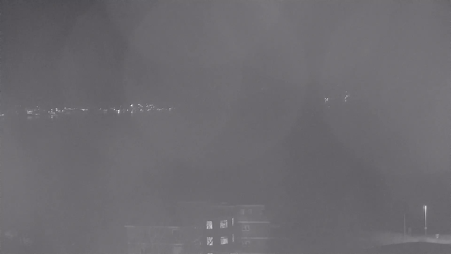 A nighttime webcam view shows a dark, cloudy atmosphere. In the distance, faint lights are visible, likely from buildings or streets along a waterfront. The foreground features a dimly lit structure, possibly a building, with some windows glowing faintly. The overall scene has a somber, hazy quality, with a significant amount of darkness and minimal detail visible.