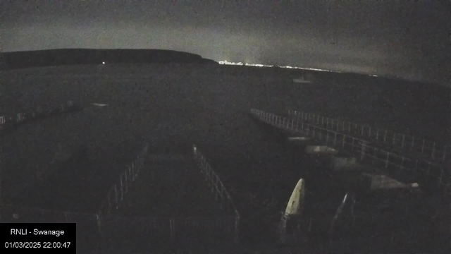 A dark image capturing a body of water with faint outlines of a pier and boats. In the background, a distant shoreline with dim lights can be seen, suggesting it is nighttime. The scene has an overall somber and quiet atmosphere. The lower left corner has a timestamp indicating the date and time of the image.