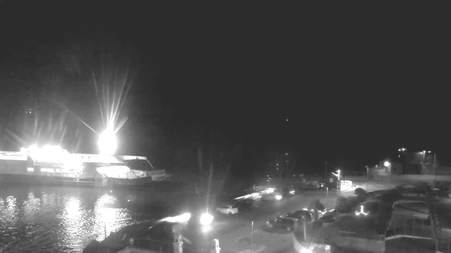 A dimly lit scene depicting a waterfront at night. A large boat is docked to the left, illuminated by bright lights on its upper deck. The reflections of the lights shimmer on the water's surface. In the foreground, there are some parked cars and streetlights, creating a faint glow in the surrounding area. The background is mostly dark with a few distant lights visible.