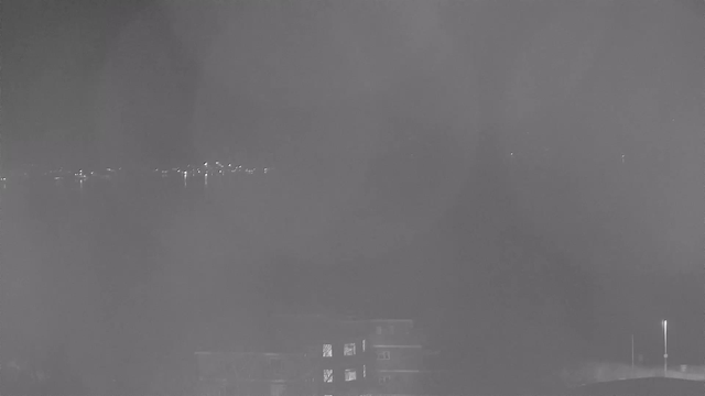 A night scene captured by a webcam, displaying a view of a calm body of water. In the background, faint lights from buildings or structures are visible along the shoreline. The image has a high contrast with a predominantly dark setting, and it appears slightly foggy or hazy, which obscures some details. In the foreground, part of a building can be seen, with faint outlines and windows reflecting some light. The overall atmosphere is tranquil and somewhat dimly lit.