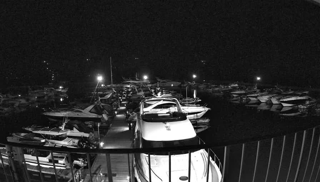 A nighttime image of a marina filled with numerous boats docked closely together. The scene is predominantly dark with faint lighting from a few poles and the reflections of boats in the water. In the foreground, a large white boat is prominent, and wooden planks can be seen as part of the dock. The atmosphere is calm and serene, with the silhouettes of boats against the night sky.