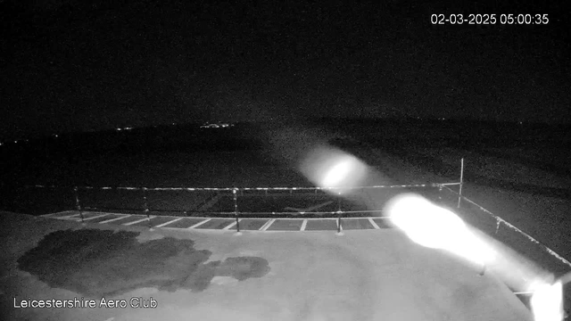 A dimly lit scene from a webcam at Leicestershire Aero Club. The image shows a flat surface, likely a runway or platform, with some dark patches on it. In the background, faint lights are visible, indicating a horizon or distant area. A barrier with vertical posts and horizontal stripes runs across the foreground. The overall atmosphere is dark, suggesting it is early morning or nighttime.