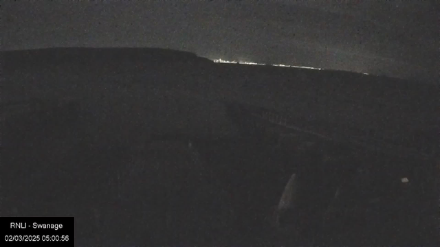 A dark nighttime scene showing a silhouette of hills or cliffs in the background. In the distance, faint lights suggest a location, possibly a town or city. The foreground is mostly obscured, with indistinct shapes that may represent rocky ground or vegetation. The overall ambiance is quiet and subdued, with minimal visibility.