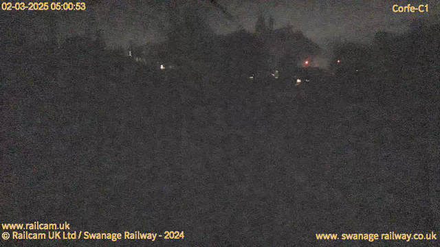 A dimly lit scene captured by a webcam. The image is predominantly dark with faint outlines of trees and structures in the background. There are small, scattered lights in the distance, possibly from buildings or vehicles. The time displayed in the top left corner shows "02-03-2025 05:00:53," indicating early morning hours. The bottom left corner includes a website link and a copyright notice, while the top right corner identifies the location as "Corfe-C1." Overall, the visibility is low, creating a nighttime atmosphere.