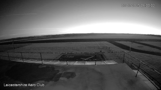 Black and white image capturing a landscape from a high vantage point. In the foreground, there's a small area with an "X" marking, possibly indicating a landing zone. Beyond this area, a grassy field stretches across the scene, bordered by a fence. The sky above is lightening as dawn approaches, with some subtle cloud patterns visible. The date and time on the image indicate it was taken early in the morning.