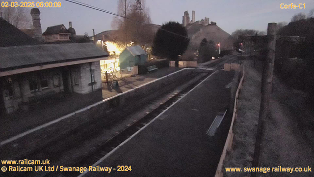 A dimly lit railway station scene at dawn. The foreground shows empty railway tracks and a platform with a wooden fence and benches. To the left, there's a building with a roof, likely the station house, partially illuminated. In the background, a hill rises with what appears to be castle ruins at the top. The sky is a gradient from dark to light, indicating the early morning. There are some trees and a slightly visible structure on the left.