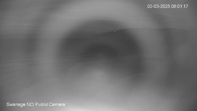 A blurry black and white image showing a faint light in the distance against an indistinct background. The scene appears foggy or low contrast, with no clear shapes or objects visible. The image includes a timestamp in the upper right corner and a label for the camera at the bottom left.