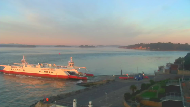 A white and red ferry is docked at a pier, surrounded by calm water reflecting soft morning light. In the background, there are green hills and a distant shoreline. Near the ferry, there are some small boats, and to the right, a landscaped area with greenery is visible. The sky has a gentle gradient of blue and soft orange hues, indicating early morning.