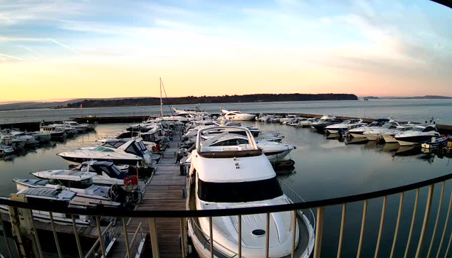 A marina scene during sunset, featuring numerous boats and yachts docked in calm waters. The sky is painted in soft pastel colors, transitioning from blue to orange and pink near the horizon. A wooden dock is visible in the foreground, and distant land can be seen on the horizon.