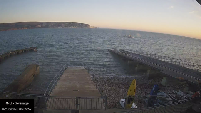 A calm seaside scene depicting a view of the water and a distant coastline. In the foreground, there are two docks, one extending into the water and the other shorter, leading to rocky shoreline. A small boat is anchored near the pier on the left. Yellow and blue kayaks are visible stored near the dock on the right. The sky is clear with a light gradient of color, indicating early morning light. The image is timestamped, showing the date and time.