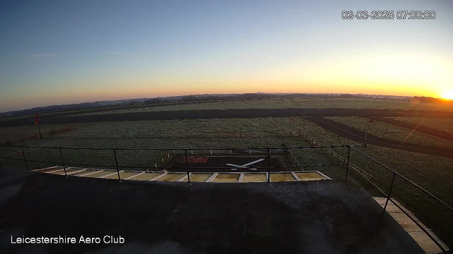 A panoramic view from a webcam at Leicestershire Aero Club showing a clear sky at dawn. The sun is rising on the horizon, casting warm tones of orange and yellow. In the foreground, there is a flat rooftop with a railing, and a small white aircraft symbol on the ground, indicating a runway. The landscape consists of green grass fields with a few distant trees, and gravel roads are visible in the distance. The image is timestamped at 07:00 on 02 March 2025.