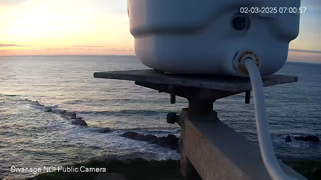 A webcam view overlooks the sea, with waves gently rolling against rocky formations. In the foreground, a white cylindrical object sits on a platform, connected by a hose. The sky is illuminated with soft hues of orange and pink as the sun rises on the horizon.