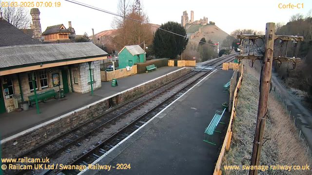 The image shows a train station platform at dawn. A wooden platform with a green bench is visible, alongside a stone building with large windows. In the background, a hill features castle ruins, and there are trees and a wooden fence nearby. The railway tracks run parallel to the platform, and a telephone pole with wires stands to the right. A sign indicating "WAY OUT" is positioned on the ground. The scene is tranquil, with soft morning light illuminating the area.