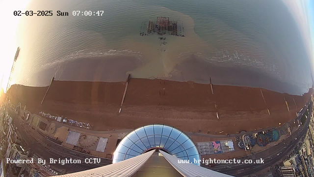 A panoramic view from a high vantage point, showcasing a sandy beach and calm water. The foreground features a circular structure with a glass dome, suggesting an observation deck. Below, the beach is lined with wooden posts extending into the water. In the distance, a partially submerged structure is visible in the water. The scene is bathed in soft morning light, with the sun just above the horizon. Various beach amenities and attractions are visible along the shore. The date and time are displayed in the upper left corner.