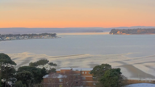 A serene view of a coastal landscape during sunset. Calm waters reflect a gradient of soft pastel colors in the sky, transitioning from orange to pale blue. Along the shoreline, small boats are anchored, and a cluster of buildings is visible, nestled among trees in the foreground. In the distance, gentle hills outline the horizon. The scene evokes a sense of tranquility and natural beauty.