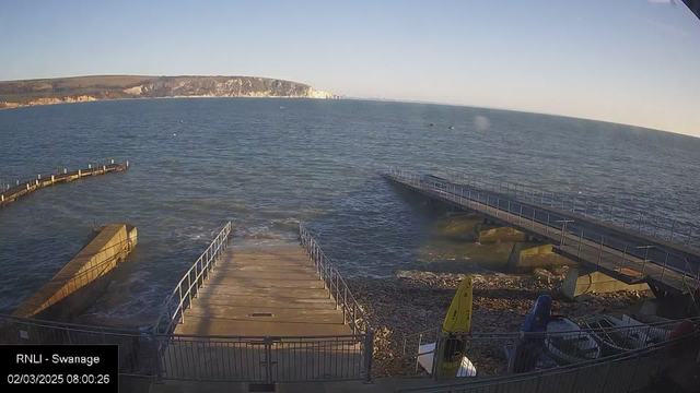 A coastal scene at Swanage, showing a calm blue sea under a clear sky. In the foreground, there are wooden piers extending into the water. To the right, a small yellow kayak is visible on the shore, next to some rocks and debris. In the background, cliffs and land are visible along the horizon. The image indicates a peaceful seaside morning.