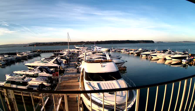 A picturesque marina scene with multiple yachts and boats moored in calm water. The foreground features a large white yacht with a sleek design, while several other boats of various sizes are docked nearby. The background showcases a clear blue sky with wispy clouds and a distant shoreline covered with trees. A wooden boardwalk extends along the marina, bordered by a railing. The tranquil water reflects the boats and sky, enhancing the serene atmosphere.