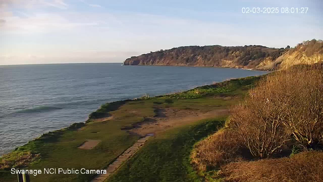 A coastal view showing the sea and a cliff in the background. The foreground features a grassy area with patches of dirt. The sky is partially cloudy with blue and white hues. A timestamp in the top right corner indicates the date and time.