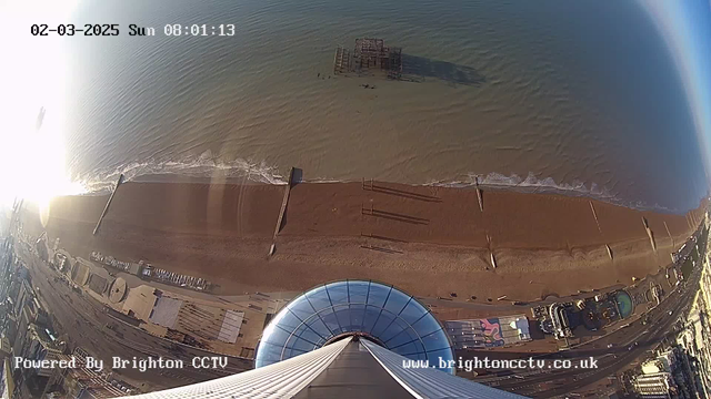 A view from a high vantage point overlooking a sandy beach and the sea. The beach is mostly empty, with gentle waves lapping at the shoreline. In the foreground, a circular structure, possibly part of an observation tower, sits at the edge. Further along the beach, there are several structures and installations, some of which appear to be amusement rides. A pier can be seen extending into the water, and there is a distant outline of a wrecked structure near the water's edge. The scene is illuminated by sunlight, creating a bright atmosphere. The image includes a timestamp at the top indicating it is 08:01:13 on March 2, 2025.