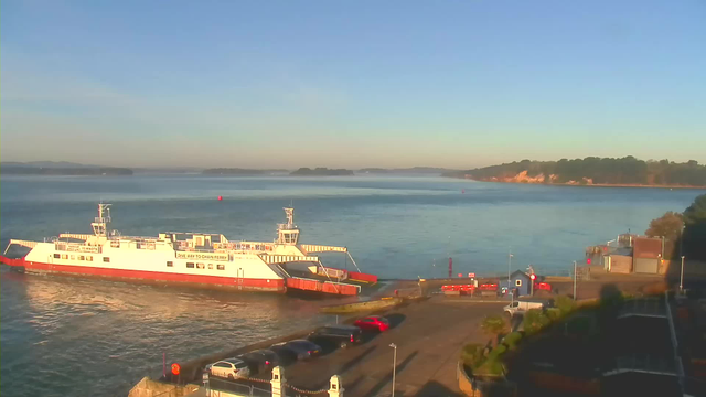 A bright morning scene at a waterfront with a large white ferry docked. The ferry has orange accents and displays a sign stating "GIVE WAY TO CHAIN FERRY." Calm water reflects the blue sky and distant hills. Vehicles are parked on the waterfront, and there are elements like lampposts and small structures nearby. The landscape includes green trees on a hill in the background, contrasting with the clear blue above.