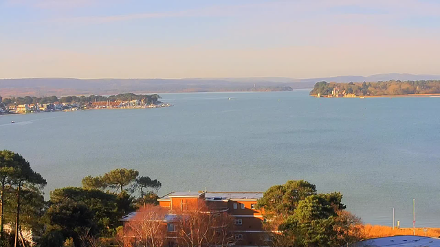 A scenic view of a calm body of water extending into the distance. On the left, there is a shoreline featuring colorful buildings and a marina filled with boats. In the foreground, trees and some buildings are visible. The background shows gentle hills and a clear sky, with soft colors indicating early morning or late afternoon light.