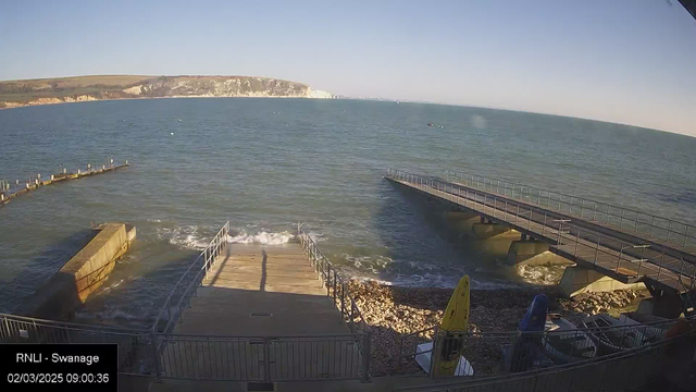 A view of the sea from a waterfront area. In the foreground, there are two wooden piers extending into the water, with one featuring stair-like steps leading down to the shore. The water is calm with gentle waves lapping against the piers. There are rocks and pebbles visible along the shore, and near the bottom right corner, there are two yellow kayaks and a blue object. In the background, white cliffs rise above the coastline under a clear blue sky. The scene is bright and sunny, indicating a clear day.