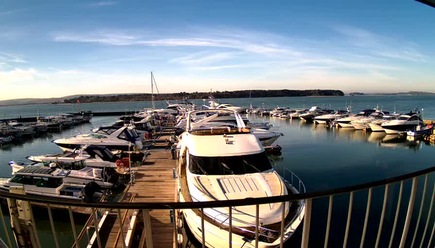 A view of a marina with numerous boats docked along wooden piers. Several white and blue yachts are visible, reflecting in the calm water. The background features a clear blue sky with soft clouds and distant hills. The scene is peaceful, capturing a sunny day at the harbor.