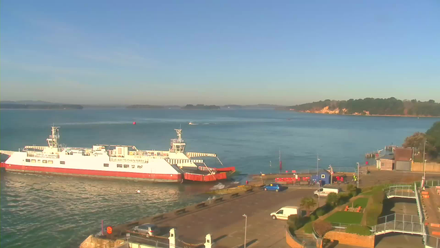 A ferry is docked at a harbor on a clear day, with calm blue water surrounding it. The ferry is white with red details and has a visible sign indicating to "give way to chain ferry." In the foreground, there is a dock with several vehicles, including a white van. To the right, there are steps leading up to a grassy area with some seating, while a small building and additional structures are visible on the right side. In the background, there are trees and a rocky shoreline along the opposite side of the water. The sky is bright blue with few clouds.