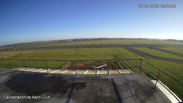 A clear blue sky is visible in the image, with no clouds present. Below, there is an expansive green field, bordered by a wooden fence. In the foreground, a small airplane is positioned on a helipad marked with a red circle and a white cross. There are paved areas in the scene, indicating taxiways or runways, with a few light traffic markings visible. The horizon features hills or distant landforms, suggesting the setting is near an airport. The timestamp on the image indicates it was taken at 9:00 AM on March 2, 2025.