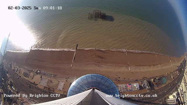 A birds-eye view of a beach and coastline, taken from a high vantage point. The image shows a sandy beach with gentle waves lapping at the shore. Reflections from sunlight create a shimmering effect on the water surface. A partially submerged pier extends into the sea, with a few vertical wooden supports visible. Beneath the viewpoint, there is a circular glass structure. Nearby, various structures and attractions are visible along the promenade, including a colorful area that appears to be an amusement park. The sky is clear and blue, indicating a sunny day.