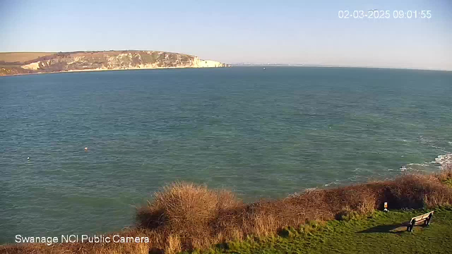 A view of the sea taken from a webcam. The foreground shows grassy land with low shrubs, while a wooden bench is positioned on the right side. The background features calm water in shades of blue and green, with a rocky cliff in the distance under a clear blue sky. The scene appears peaceful and serene, capturing a bright and sunny day.