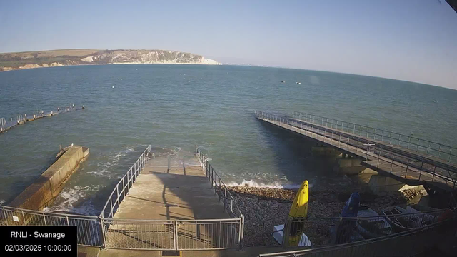 A view of the sea with gentle waves and a clear blue sky. In the foreground, there is a concrete ramp leading down to the water, flanked by metal railings. To the left, a low, walled dock extends into the water, while to the right, a wider pier with a railing is visible. Several boats are moored nearby, and a yellow kayak is positioned on the shore. The distant cliffs in the background show a mix of land and rock formations.