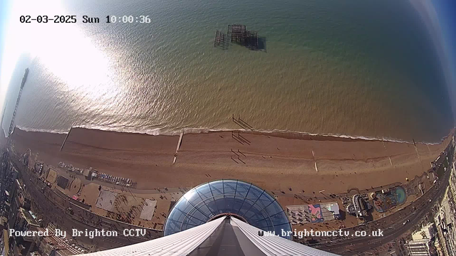 A high-angle view of a beach on a sunny day. The coastline features a long stretch of golden sand, with gentle waves lapping at the shore. Various structures line the beach, including a pier and amusement rides. In the background, the ocean transitions from blue to green hues, reflecting sunlight. A transparent dome structure is visible in the foreground, contributing to the beach scene. The image includes a timestamp and the text indicating it is powered by Brighton CCTV.