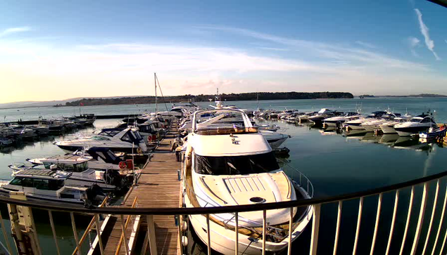 A marina scene showing several boats docked at a wooden pier. The foreground features a large white yacht with a brown stripe and a sunbathing area. The water is calm and reflects the light from a clear blue sky, with slight clouds in the background. Other boats, varying in size and color, are lined up along the pier, surrounded by a serene landscape of distant hills.