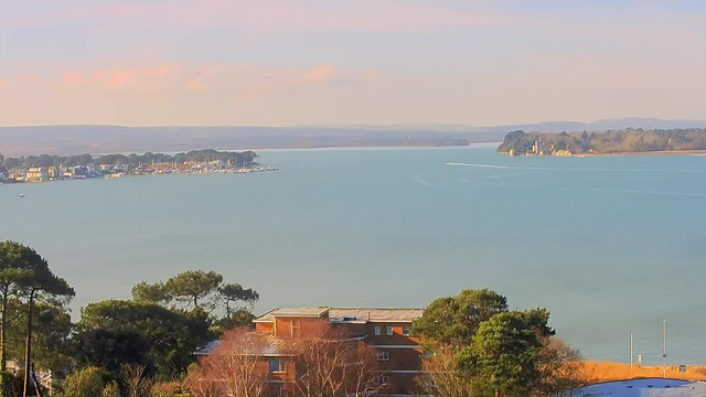 A scenic view of a calm bay or body of water, with a small town visible along the shoreline. The water appears tranquil and reflects soft shades of blue. In the foreground, there are trees and buildings, including a red-brick structure. There are several boats scattered in the water, and in the distance, a small peninsula or island with greenery is visible. The sky contains light clouds and a gentle color gradient indicating either early morning or late afternoon light.