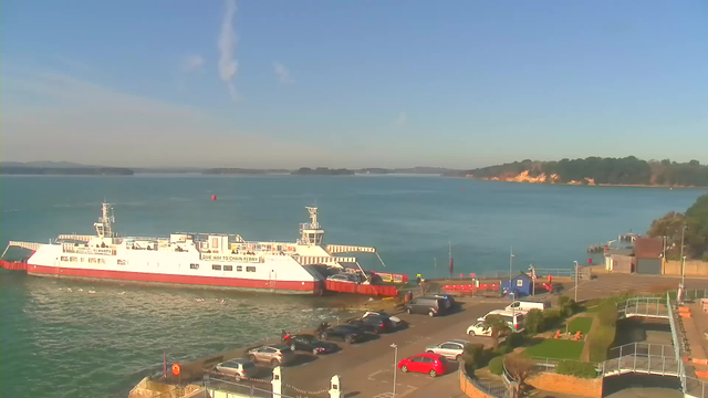 A white ferry with a red stripe is docked on a blue body of water. The sky is clear with a few wispy clouds. In the foreground, there is a parking area with several cars and a pathway leading to a seaside area. On the right side, there are buildings and greenery. In the background, you can see a coastline with trees and a hint of distant land in the water.