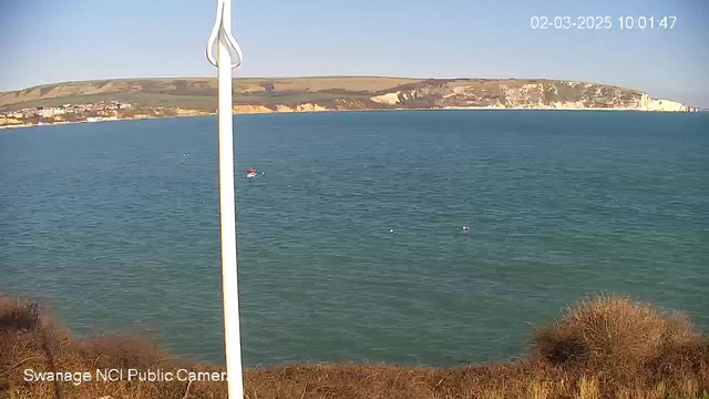 A coastal scene showing calm turquoise water under a clear blue sky. In the foreground, there is a white pole, possibly a marker or flag. The water features a few small boats, one of which has a red and white hull. The background includes green hills leading up to a rocky cliff, with a small town visible on the shore. The image is bright and sunny.