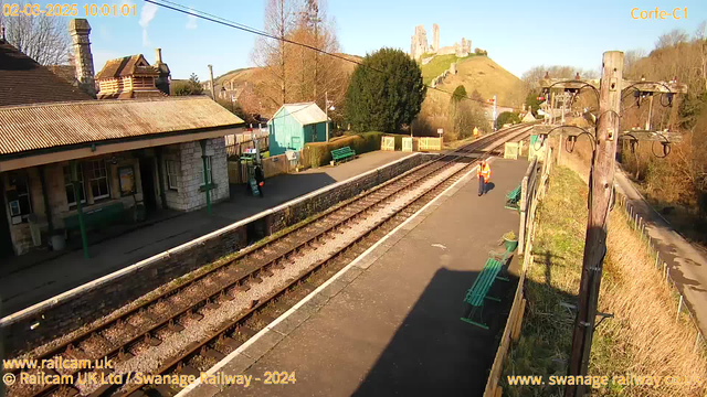 A sunny train station scene with a platform lined by railway tracks. A person wearing an orange safety vest is walking on the platform. In the background, there is a stone building with a sloped roof, and nearby, a small green shed and several green benches are visible. Beyond the station, a hill rises with a castle ruin at the top. The sky is clear with a few clouds, and there are trees on either side of the platform.